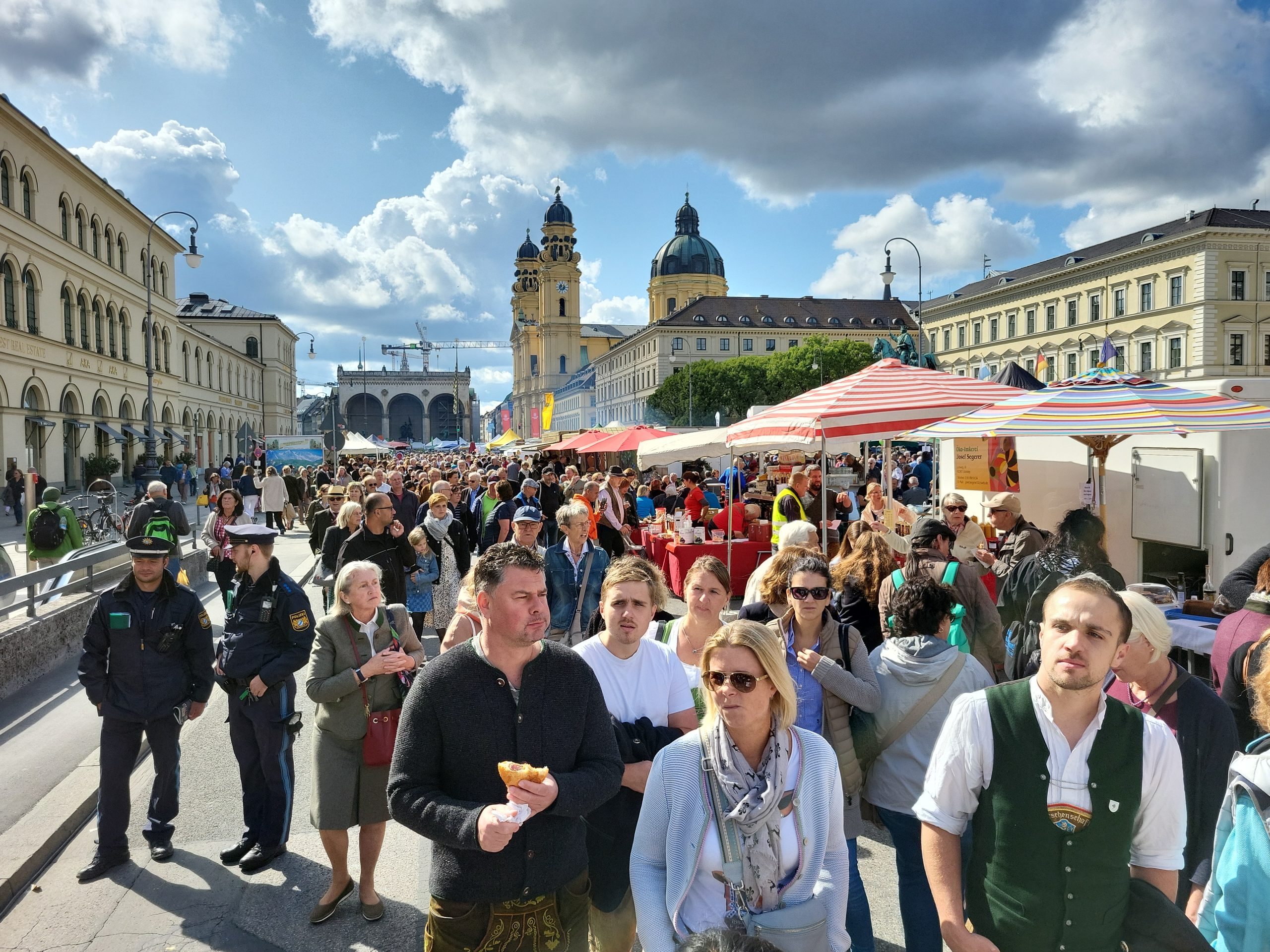 Bauernmarktmeile_c_BBV (3) - IN München - Das Stadtmagazin Crowded outdoor street market with colorful stalls and striped tents, set against historic buildings and domed towers in the background.