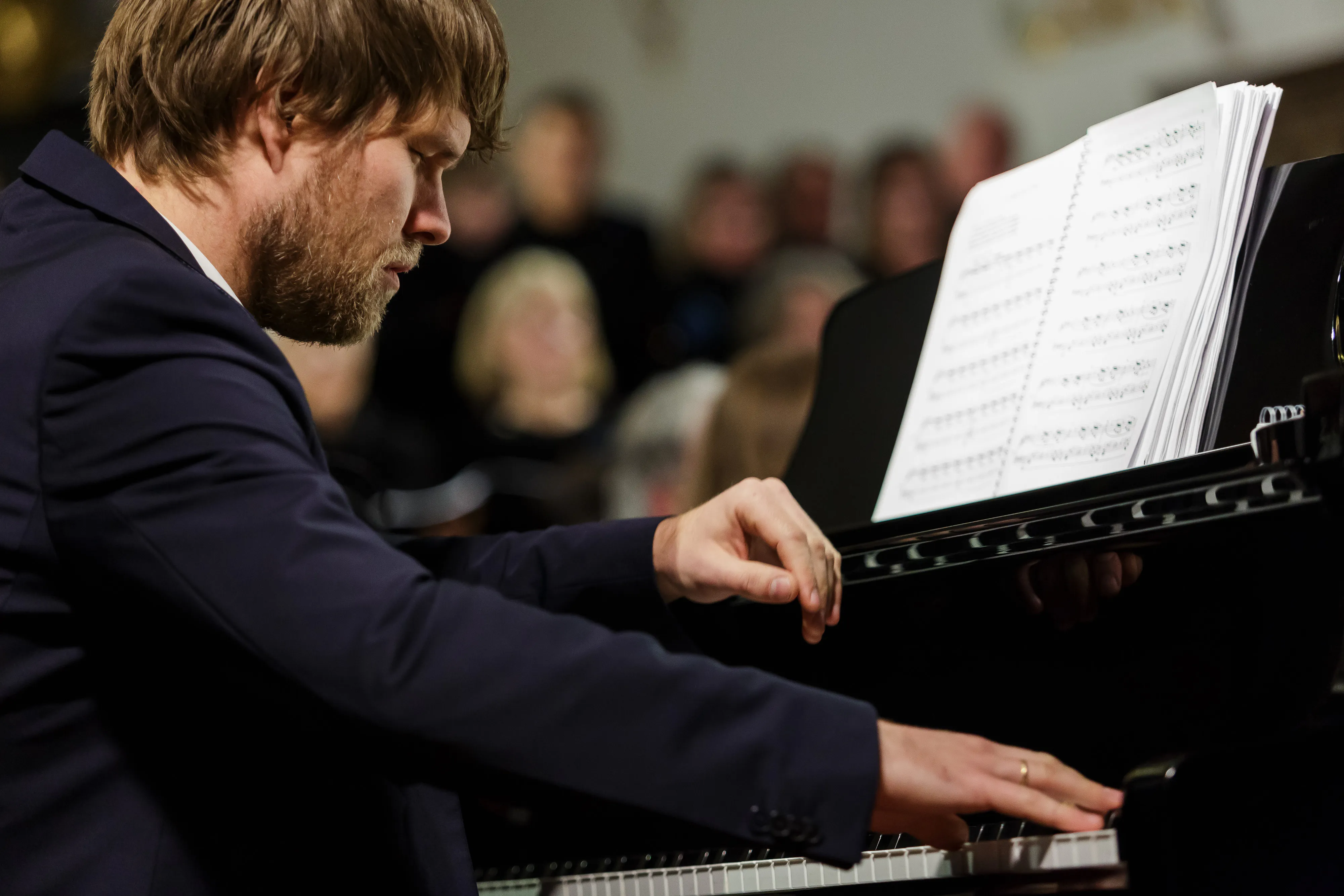 Begert_TobiasGutmann - IN München - Das Stadtmagazin Pianist in a dark blazer focused on playing a grand piano, sheet music visible on the stand with an audience blurred in the background.