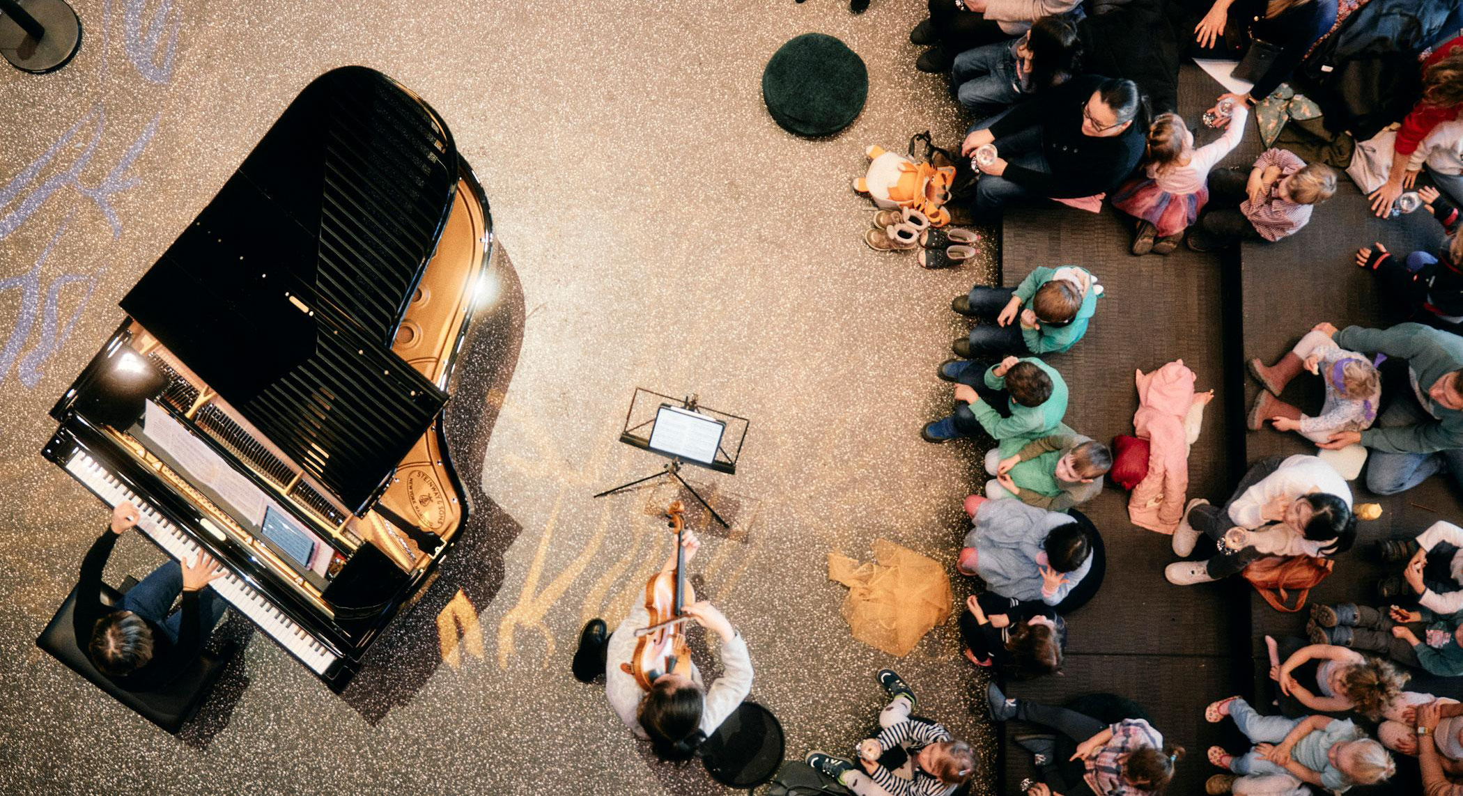 Overhead view of a black grand piano on a glittery stage with a pianist, a cellist nearby, and a crowd seated and standing around the performance area.