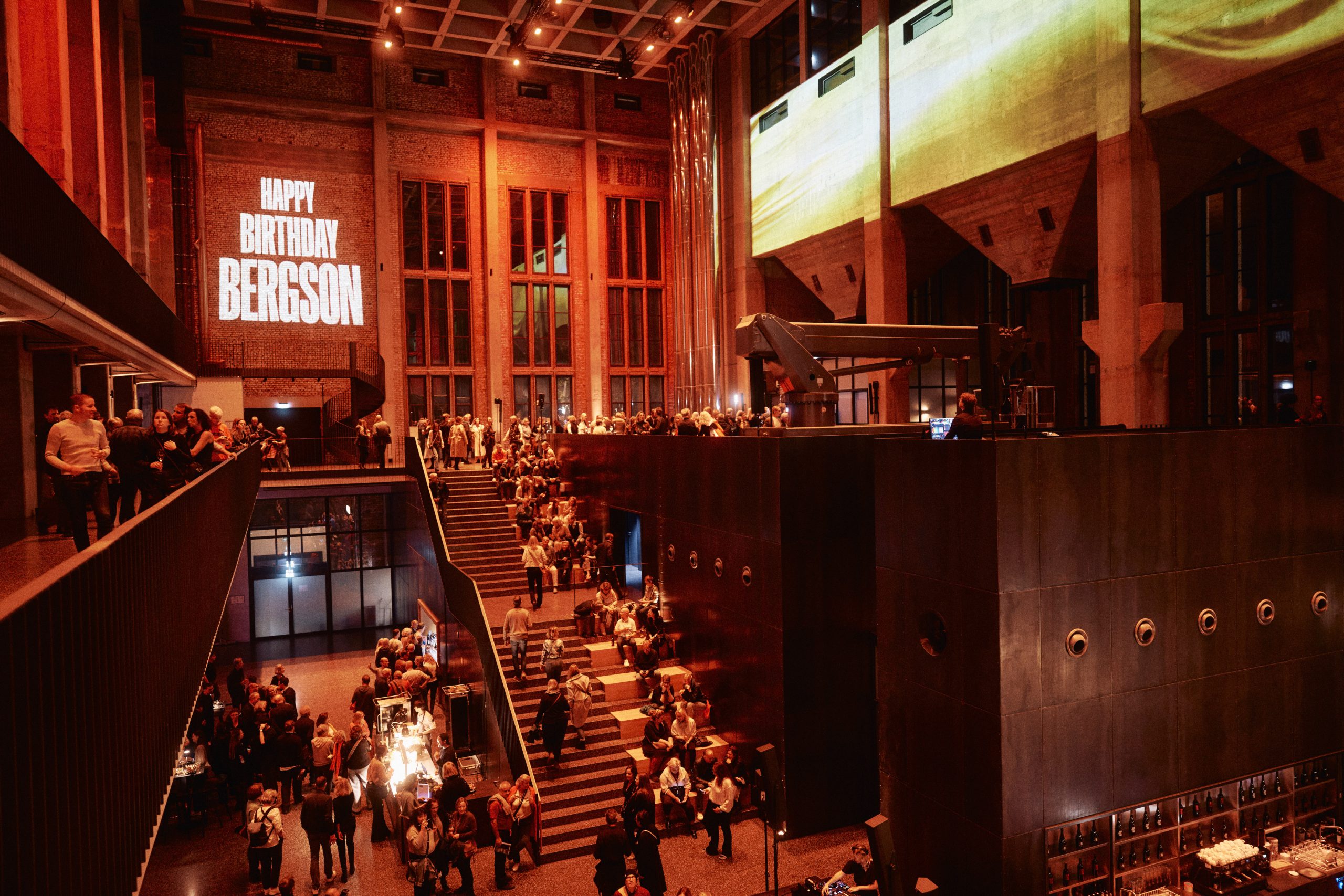 Bergson_bySimonHaseneder - IN München - Das Stadtmagazin Large indoor venue with red lighting, crowd on stairs and balconies; wall reads'HAPPY BIRTHDAY BERGSSON'.