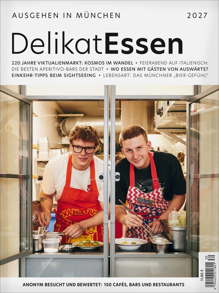 001_Deli__TITEL 2026_27_ohneRuecken.indd - IN München - Das Stadtmagazin Two men in red aprons stand behind a glass kitchen window, cooking and plating food together.