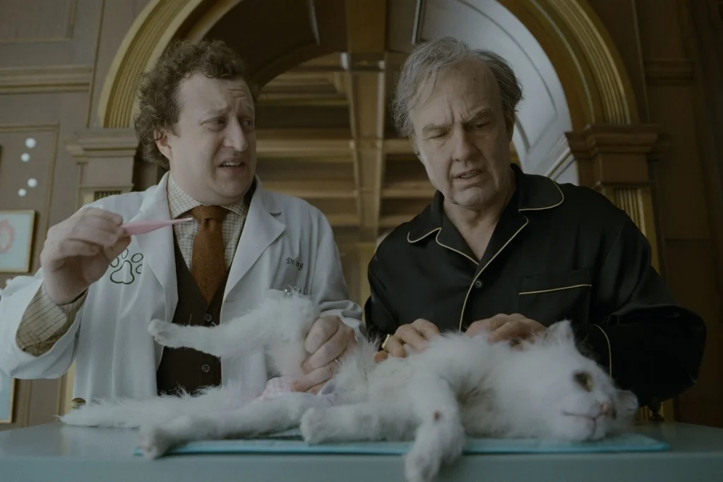 Digger - IN München - Das Stadtmagazin Two men in a lab coat and pajamas examine a white fluffy cat on an examination table in a warm, wood-paneled room.