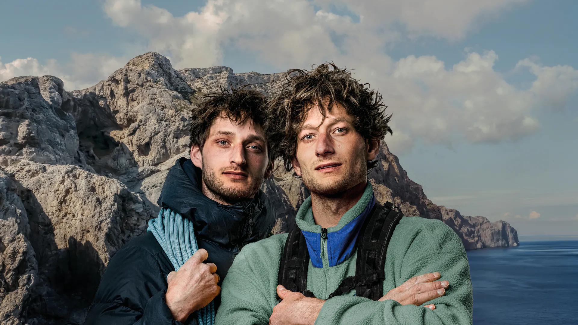 Two friends posing arm-in-arm on a rocky coastline with the sea in the background, rugged cliffs behind them.