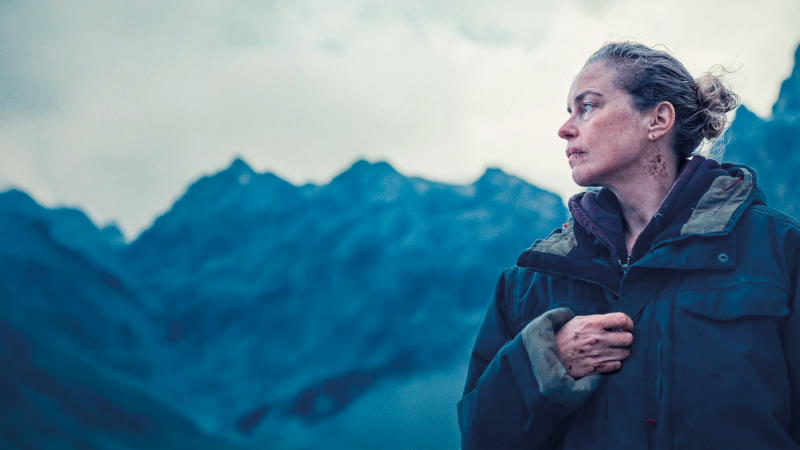 Woman in a dark jacket stands on a misty mountain ridge, looking off to the right with a somber expression.