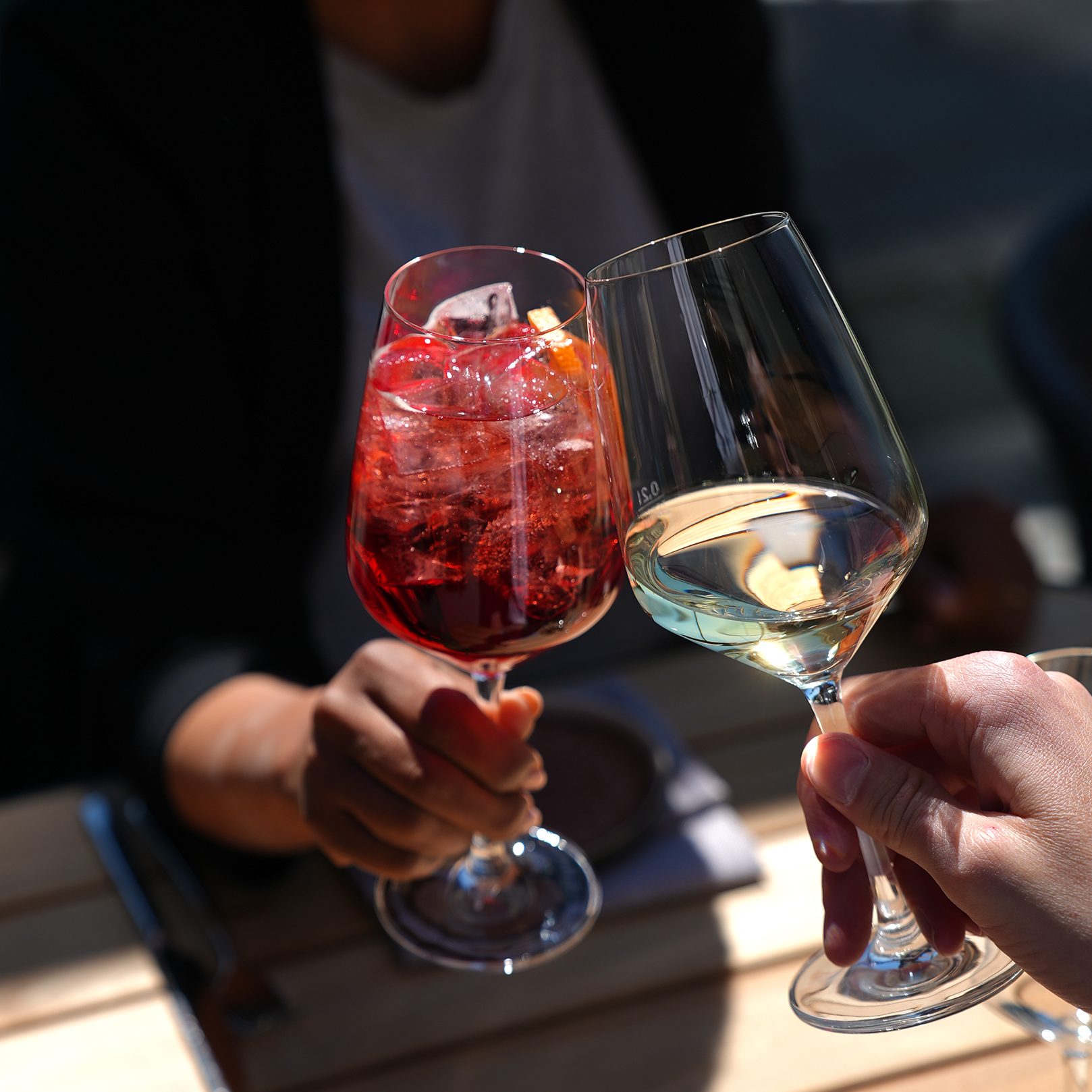 Friends raise glasses in a toast; a red cocktail with ice and a white wine glass visible outdoors.