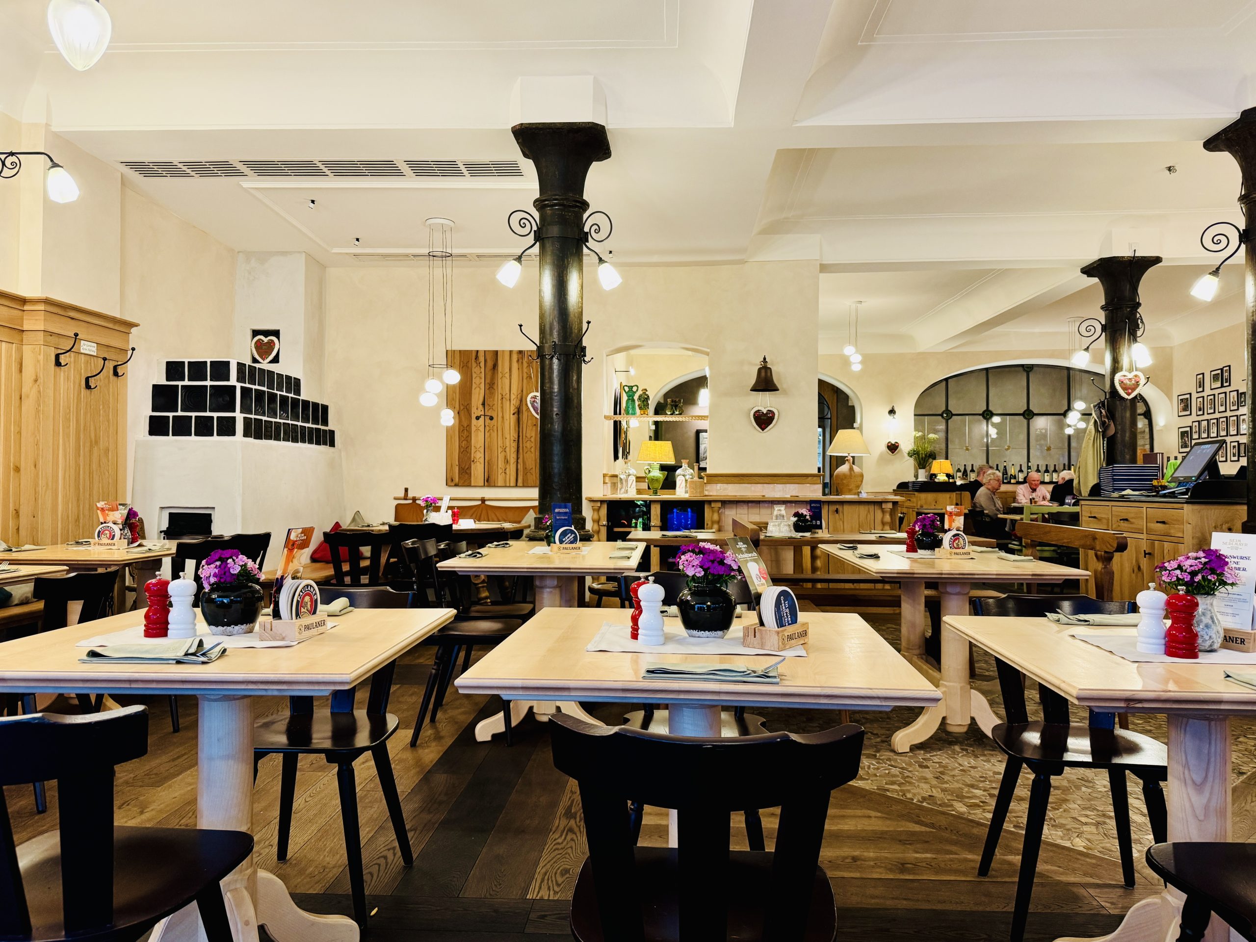 Bright cafe interior with wooden tables and black chairs, a central dark pillar, and warm pendant lights.