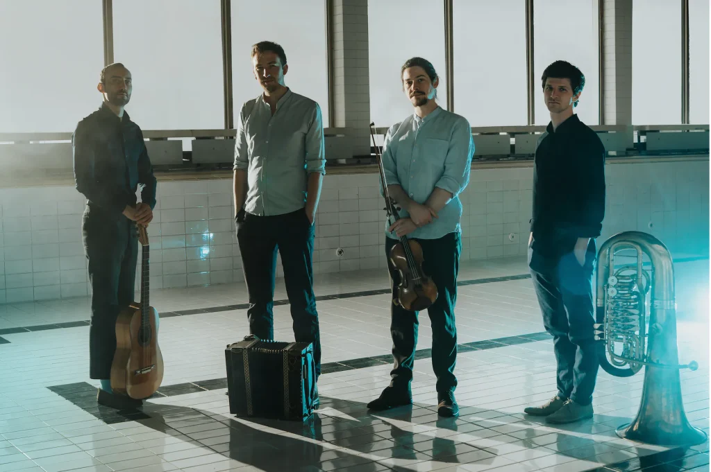 Maxjoseph_quer_JessicaElsner - IN München - Das Stadtmagazin Four men stand in a tiled pool area with their instruments: guitar on the left, a violin held by the third man, an accordion on the floor, and a tuba nearby on the right.