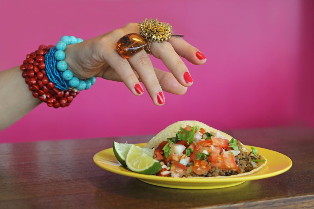 Hand with red nails and colorful bracelets hovering over a taco on a yellow plate with lime wedges.