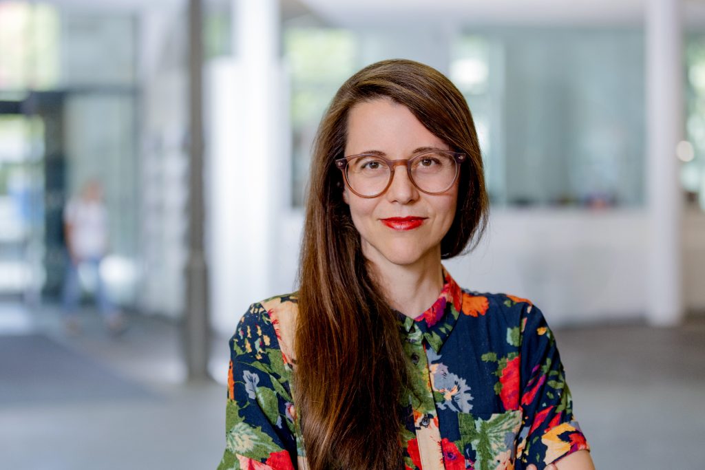 2025_07_23_FotoLoredanaLaRocca_1185 - IN München - Das Stadtmagazin Portrait of a woman with long brown hair, wearing round glasses and a colorful floral shirt, in a bright indoor hallway.