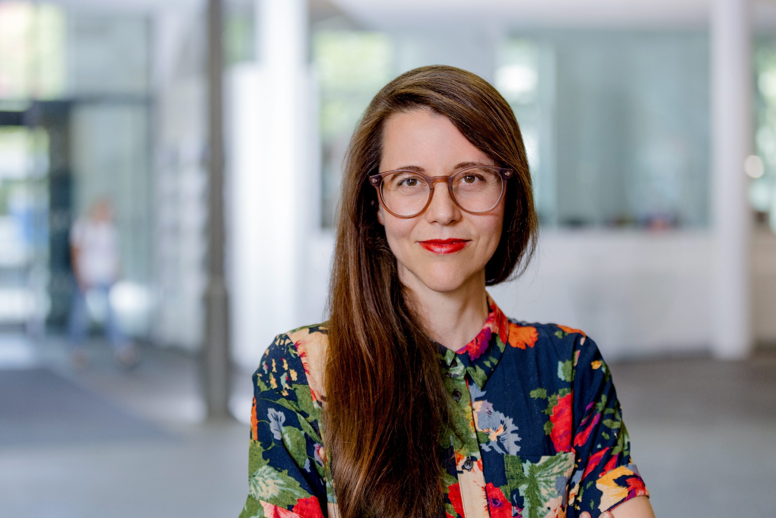 Portrait of a woman with long brown hair, wearing round glasses and a colorful floral shirt, in a bright indoor hallway.