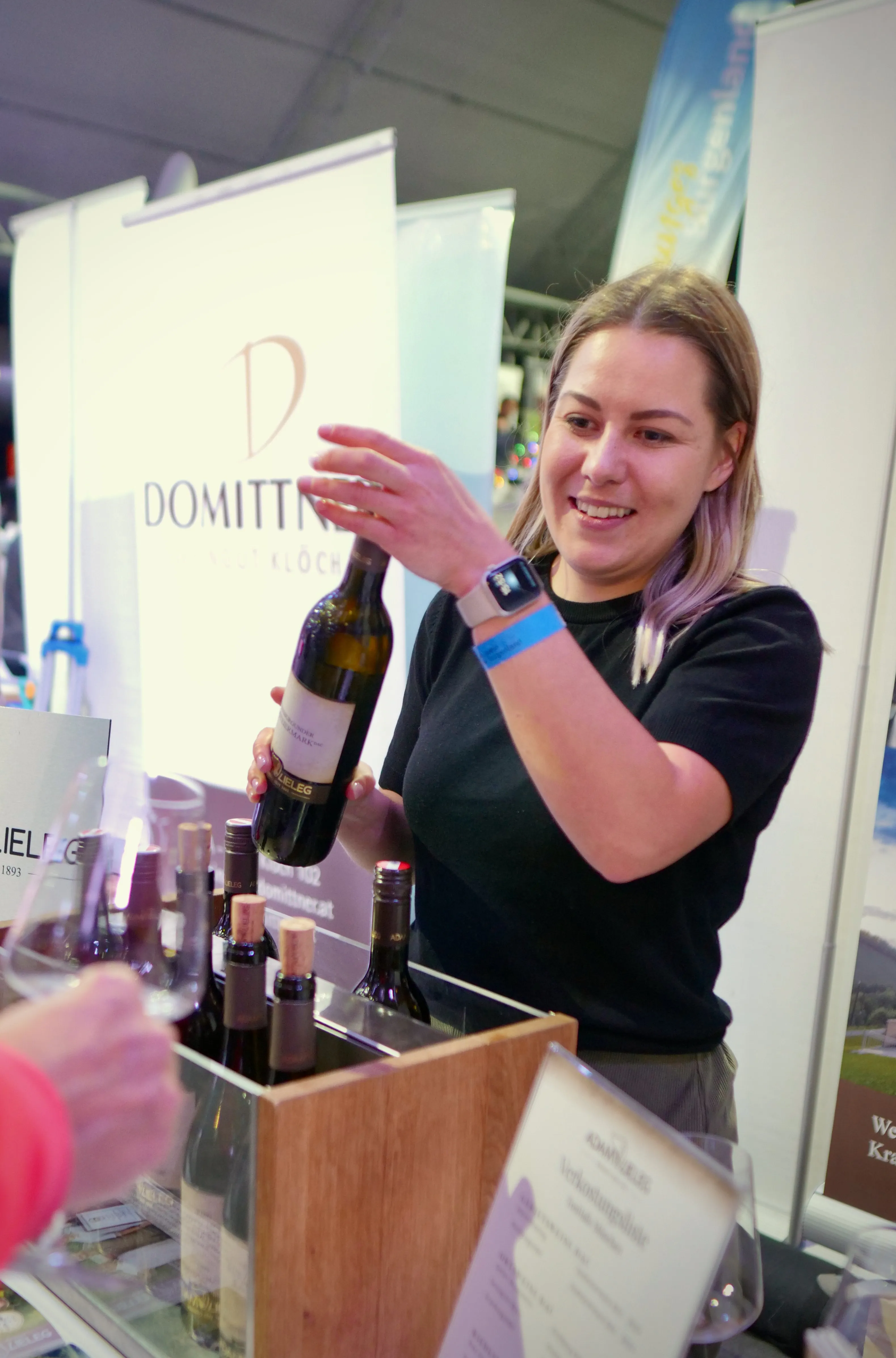 Smiling woman at a booth pouring wine from a bottle into a glass, with several bottles lined up on the counter in front of her.