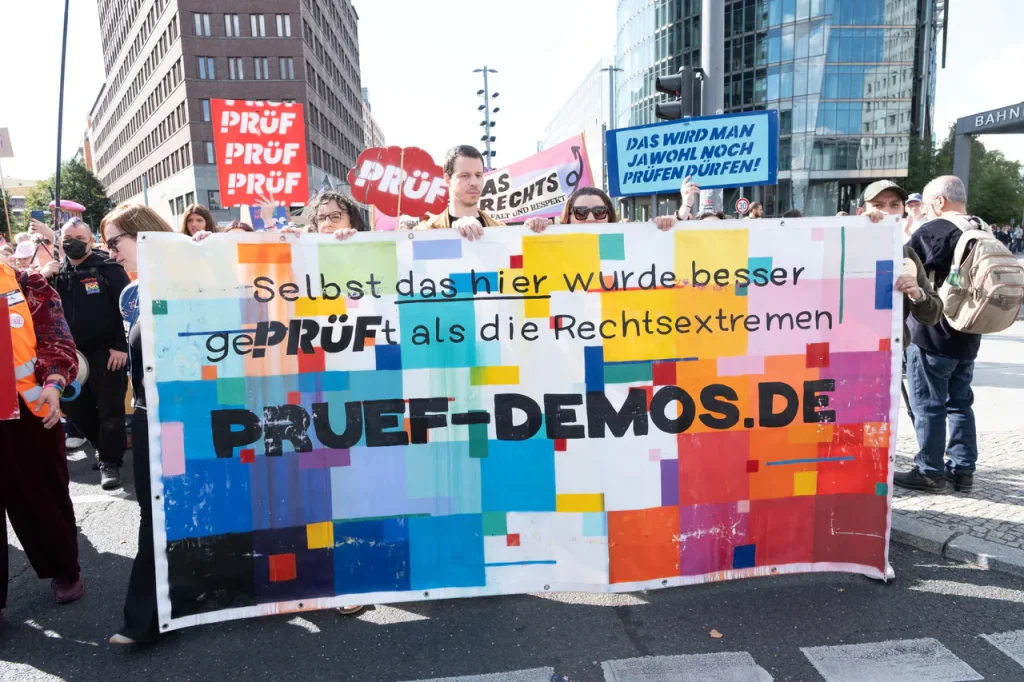 Protesters hold a colorful banner reading 'PRÜF-DEMOS.DE' in front of a city street, with signs in German in the background.
