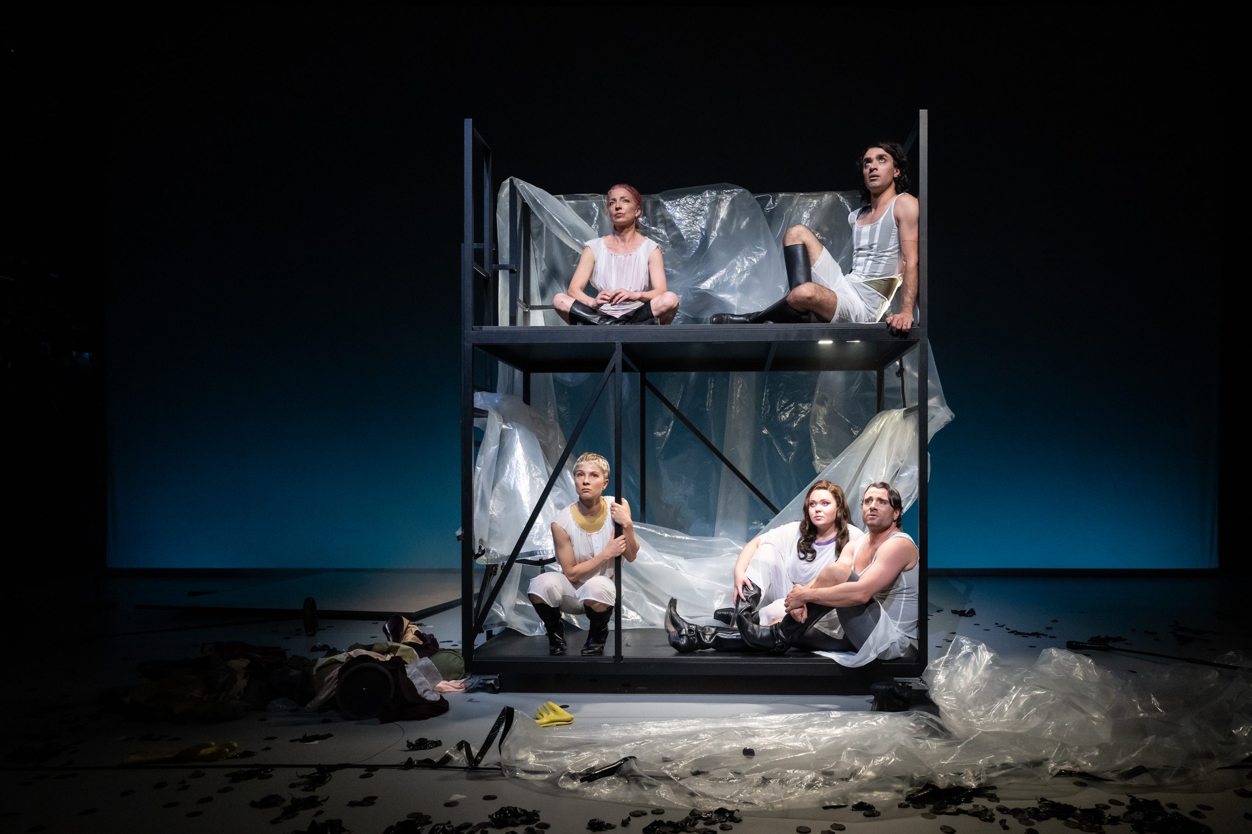 Resi_Reigen_-_Nicola_Kirsch__Pujan_Sadri__oben___Lea_Ruckpaul__Alexandra_Juschkewitsch__Florian_Jahr__c__Sandra_Then - IN München - Das Stadtmagazin Actors sit on a metal platform wrapped in clear plastic, performing on a dark stage with blue gradient floor lights behind.