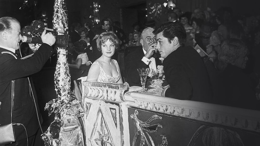RomySchneider_AlainDelon - IN München - Das Stadtmagazin Black-and-white photo of a formal banquet: a cameraman films a couple seated at an ornate table while guests watch nearby.