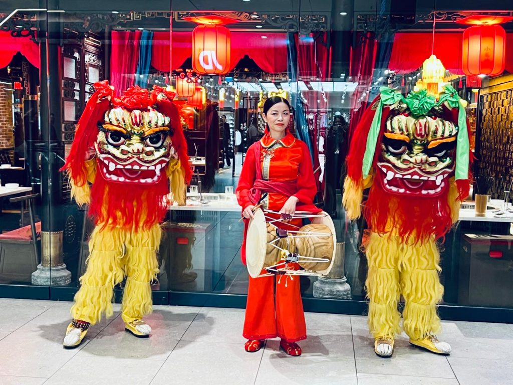 Shoo_Loong_Kan_Hotpot_Neueroffnung - IN München - Das Stadtmagazin Two colorful lion dance performers stand beside a woman in a red traditional dress playing a drum in a shop window setting for a celebration.