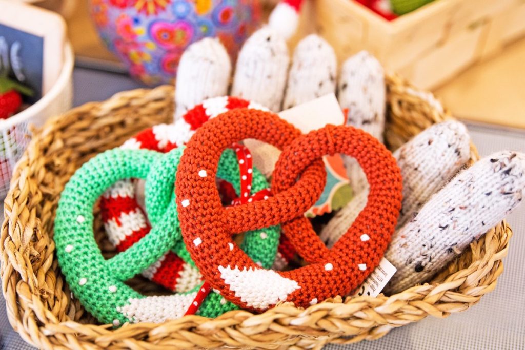 Knitted red and green crochet loops (pretzel shapes) with white polka dots in a wicker basket, colorful yarn items nearby.