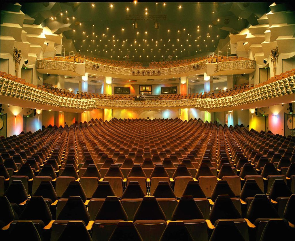 Theatersaal_nach_Sanierung_1982_Foto_Deutsches-Theater - IN München - Das Stadtmagazin Interior of a large theater with many brown seats and multiple balconies, softly lit overhead.