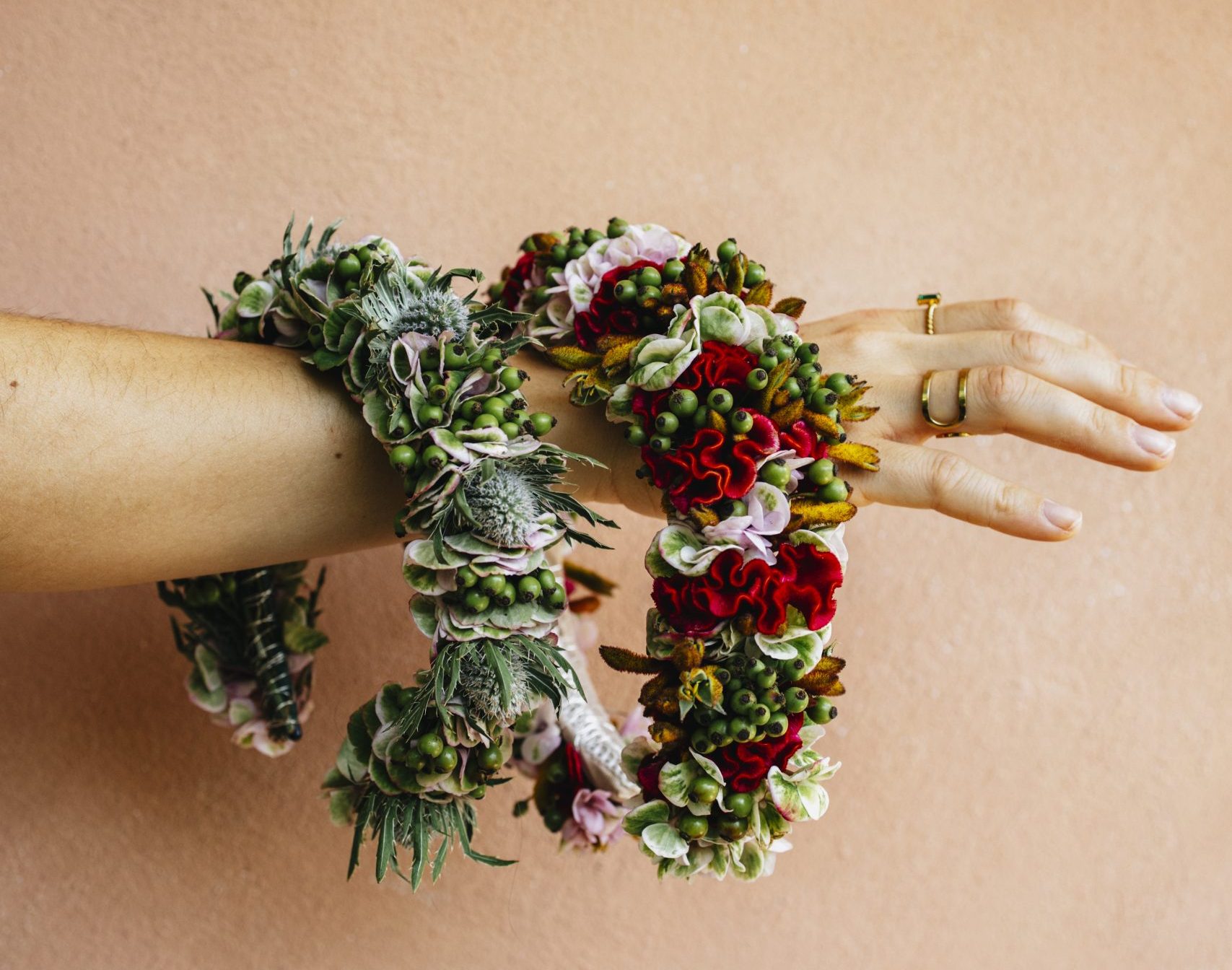 Person's forearm adorned with a colorful floral garland, featuring red, pink, and green blooms and berries, while wearing rings on the fingers against a peach wall.