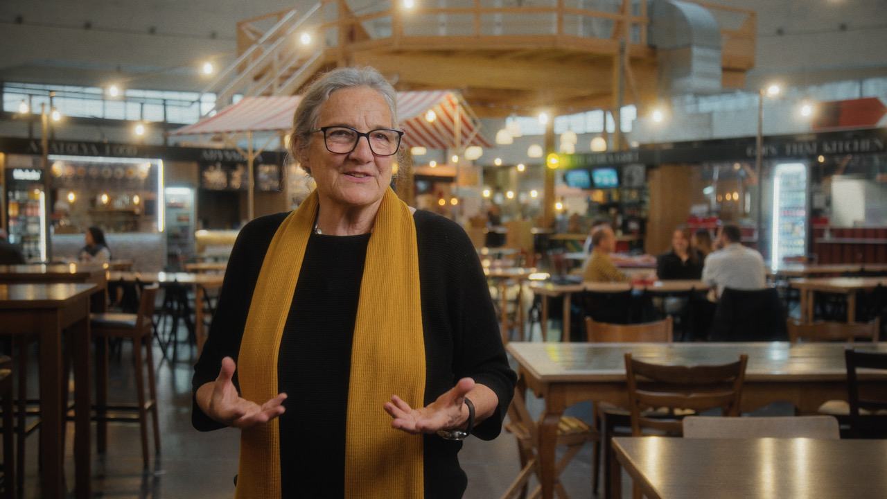 Senior woman with glasses and a mustard scarf speaks with her hands raised in a busy indoor market or food hall.