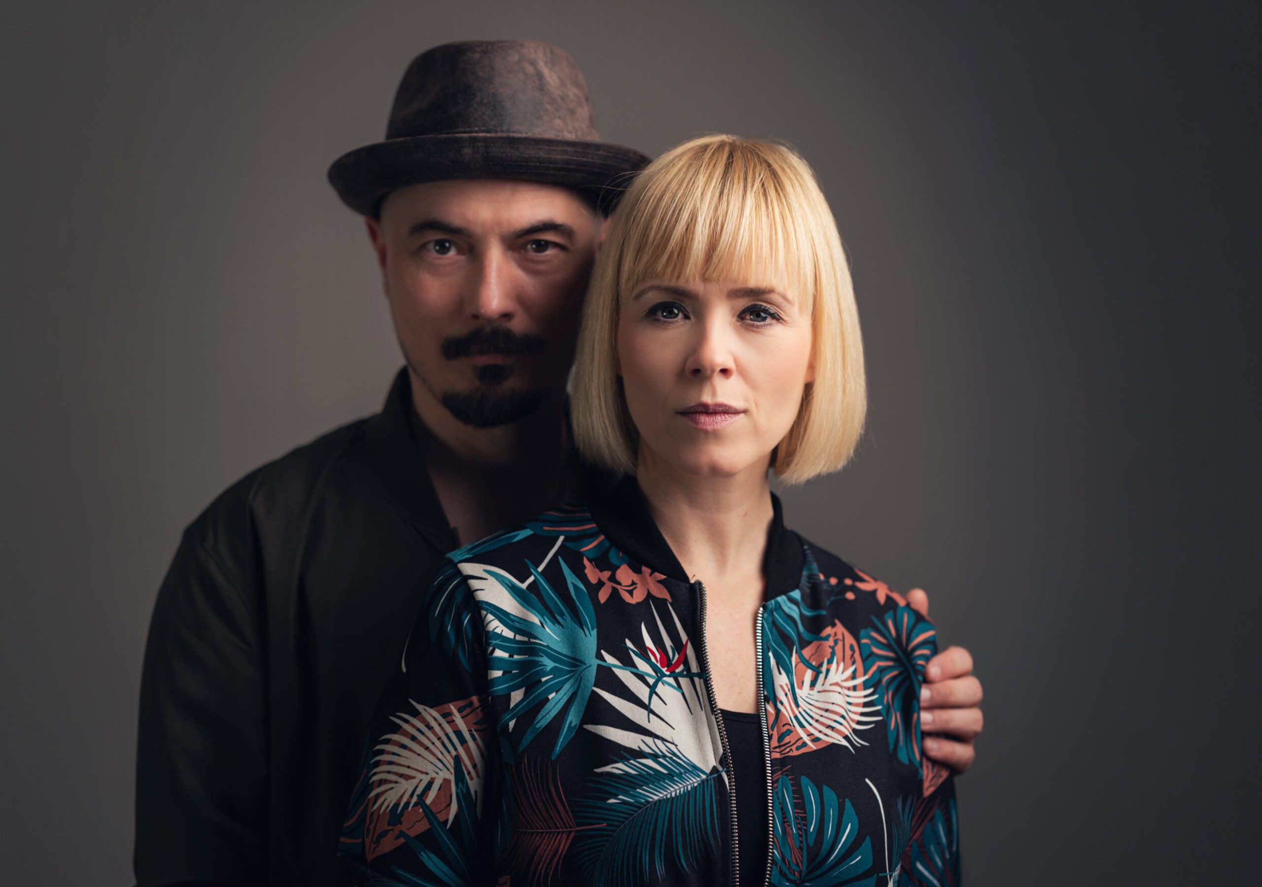 Close-up of a blonde woman in front, with a man wearing a hat standing behind her, both looking at the camera.