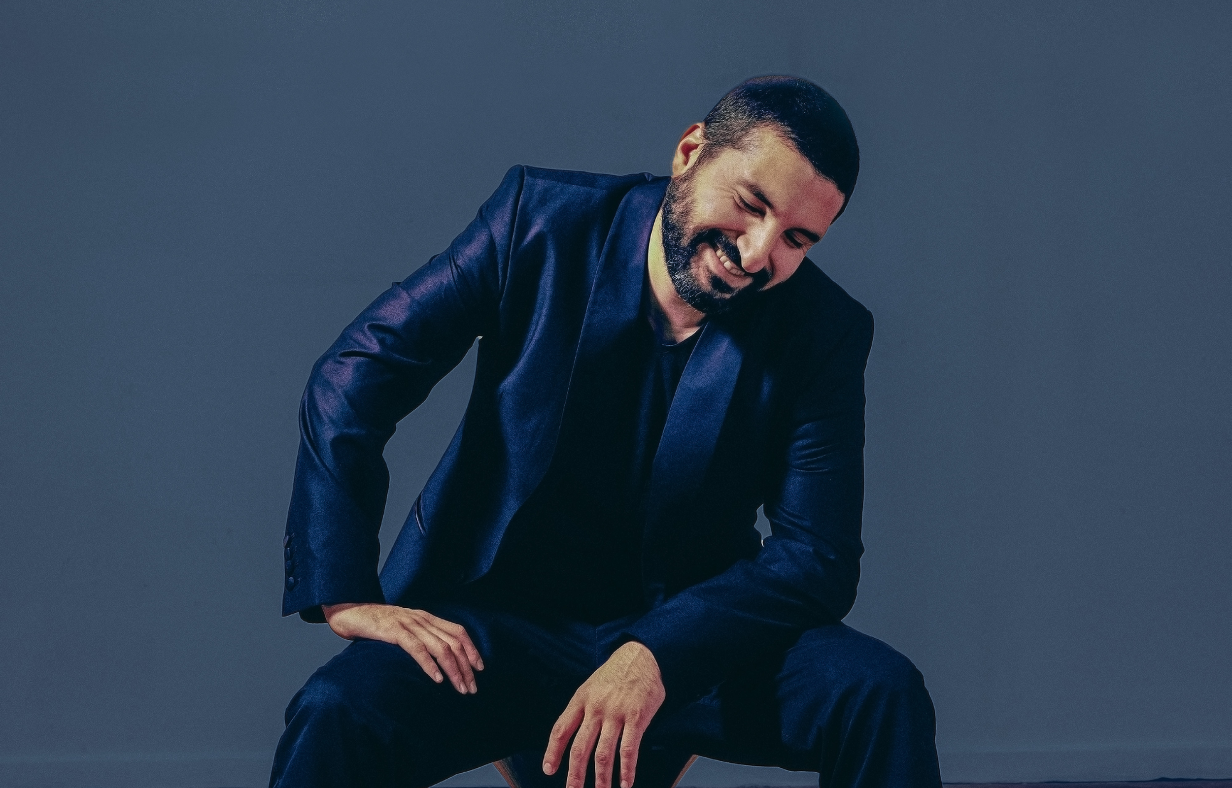 Man in a navy suit sitting and smiling, looking down with a relaxed posture against a dark blue backdrop.