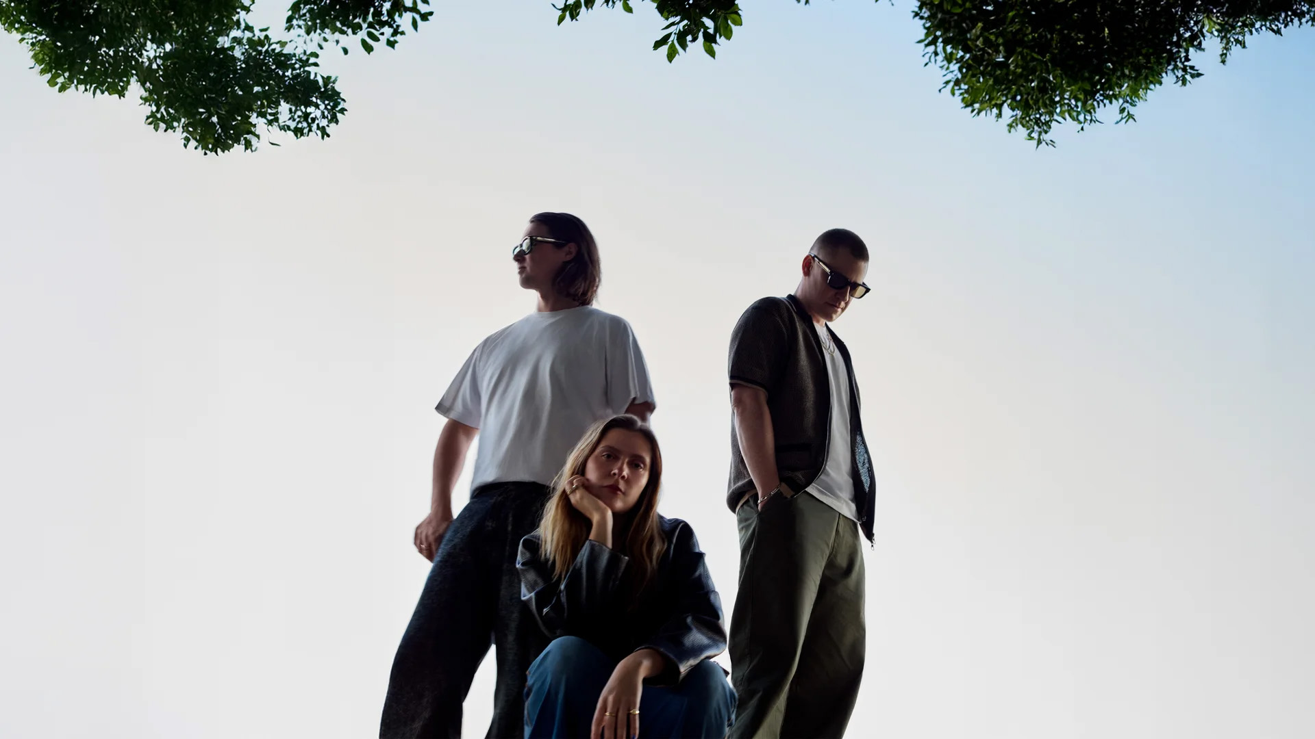 Three friends posing outdoors against a pale sky, with tree branches visible above; two standing, one seated in front.