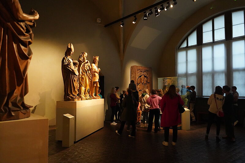 Museum gallery with visitors viewing large stone statues on pedestals by a barred arched window.