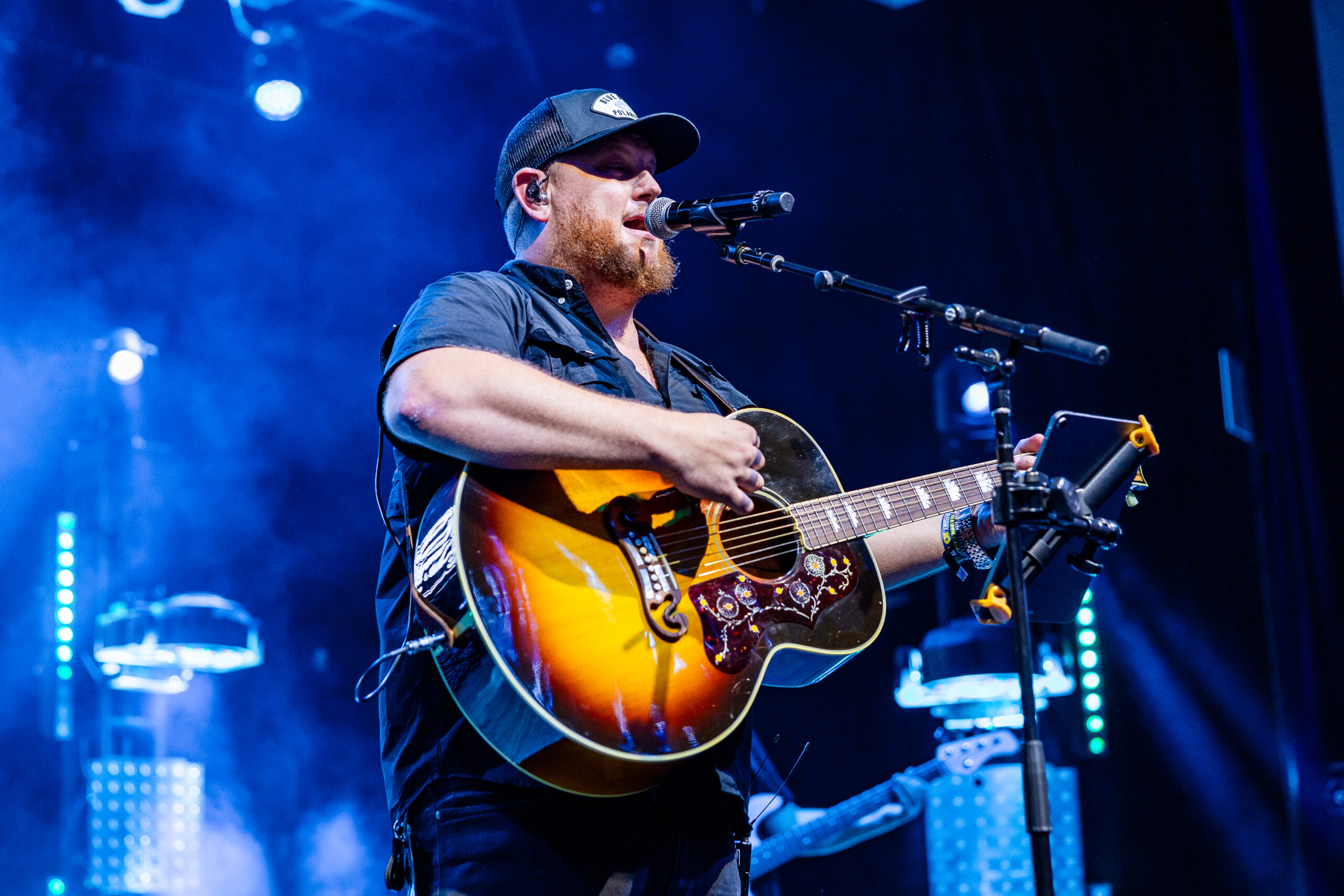 Male guitarist with beard and cap singing into a microphone on a blue-lit stage during a live performance by the guitarist on stage with an acoustic-electric guitar.