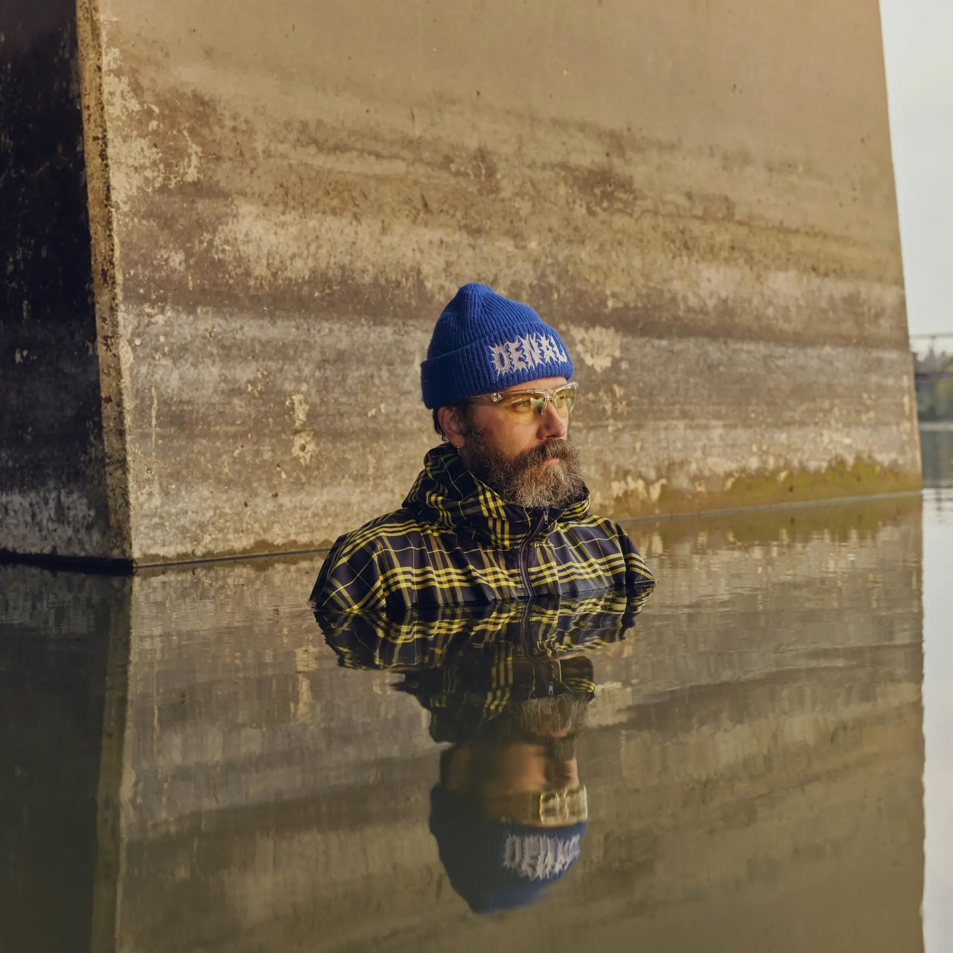 Man wearing a blue knit beanie and plaid jacket stands near a weathered stone wall, with his reflection visible in shallow water.