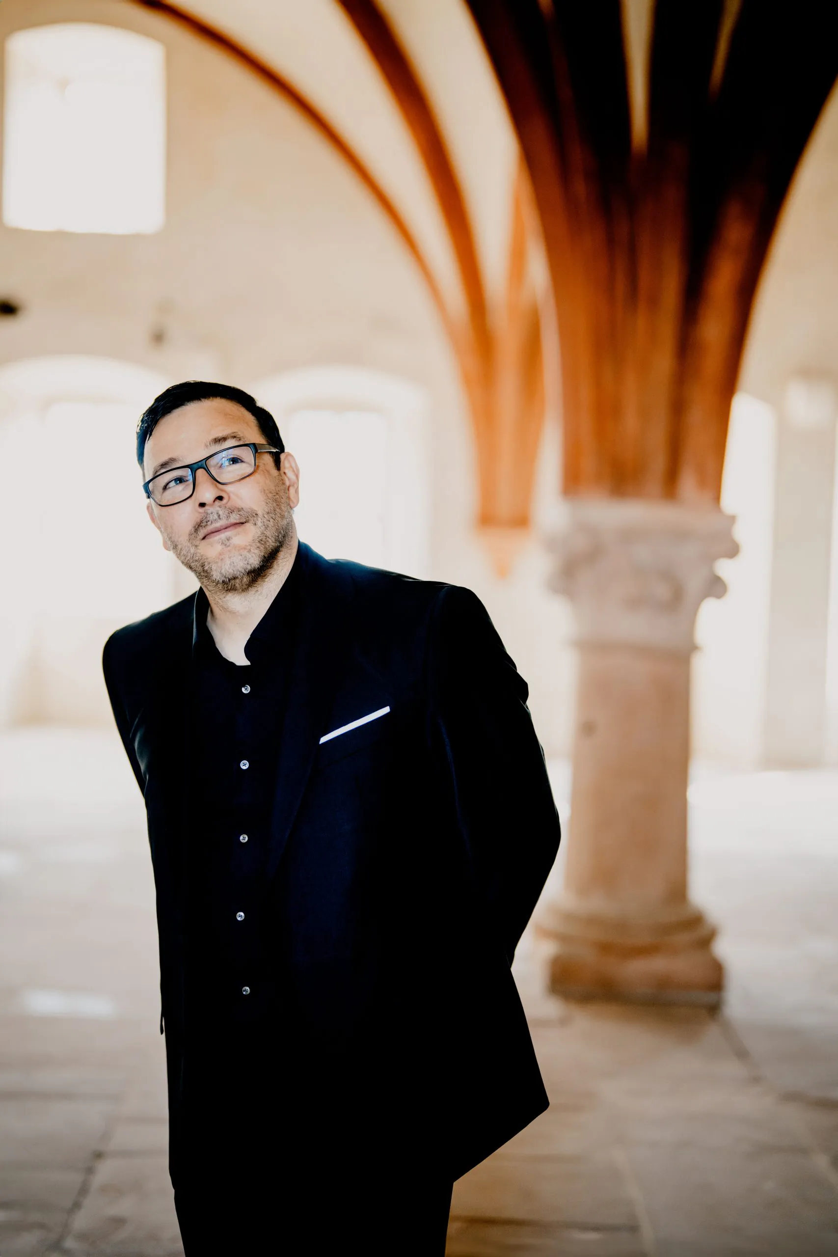Portrait of a man wearing glasses and a black suit in a sunlit arched hallway.