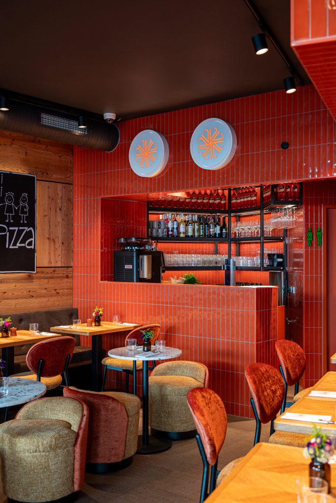 Restaurant bar area with red tiled walls, round blue signs with an orange floral logo, and glassware behind the counter.