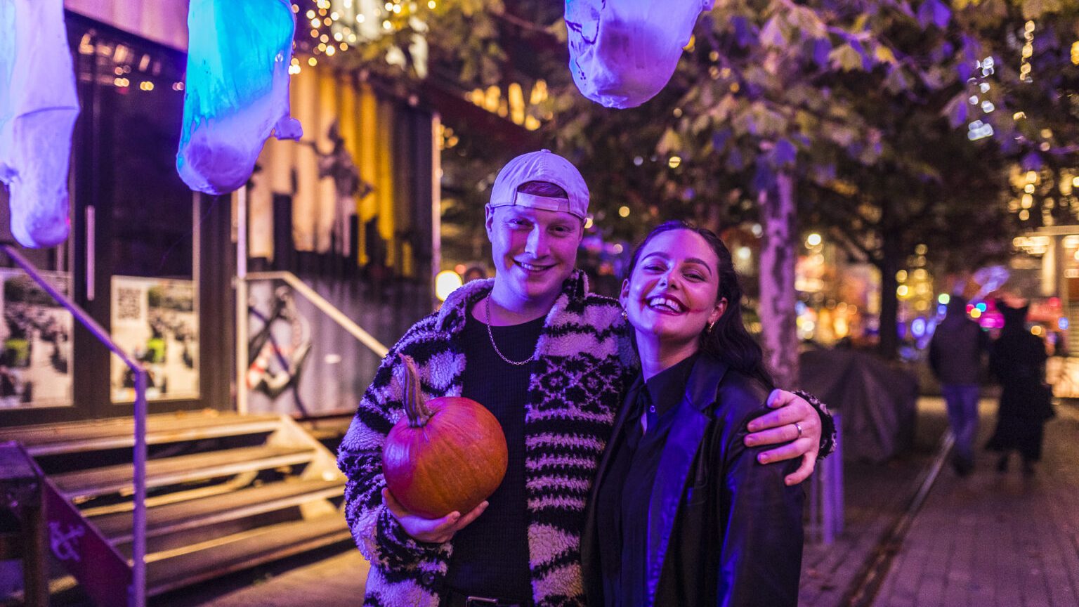 Werksviertel_Mitte_Halloween_2026 - IN München - Das Stadtmagazin Smiling couple posing with a pumpkin at a nighttime street festival, purple lighting and decorations overhead.