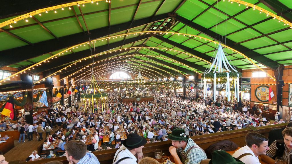 Augustiner Festhalle © München Tourismus Tommy Loesch - IN München - Das Stadtmagazin Crowded beer hall with long communal tables, festive banners, and string lights inside a green-roofed venue.