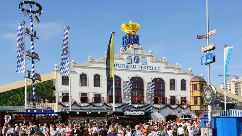 Aussenansicht Hofbraeu Festzelt © München Tourismus Tommy Loesch - IN München - Das Stadtmagazin Hofbräu Festzelt beer hall with blue-and-white banners and a crowd at a Bavarian festival.