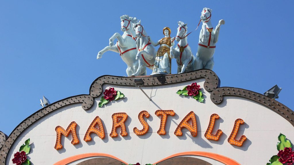 Marstall Wiesnfestzelt Aussenansicht 3 © München Tourismus Tommy Loesch - IN München - Das Stadtmagazin Ornate carnival marquee with a statue of a gold-clad woman riding a white horse-drawn wagon on top, set against a blue sky.