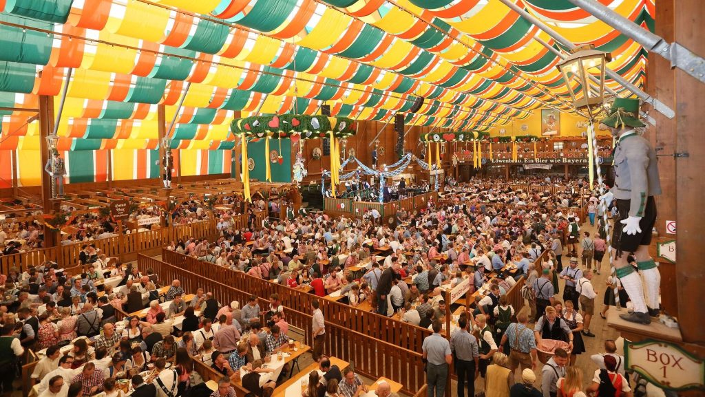 Schuetzen-Festzelt, © München Tourismus, Tommy Loesch - IN München - Das Stadtmagazin Crowded beer hall with long wooden tables under a colorful orange, yellow, and green canopy, people enjoying food and drinks in traditional Bavarian attire.