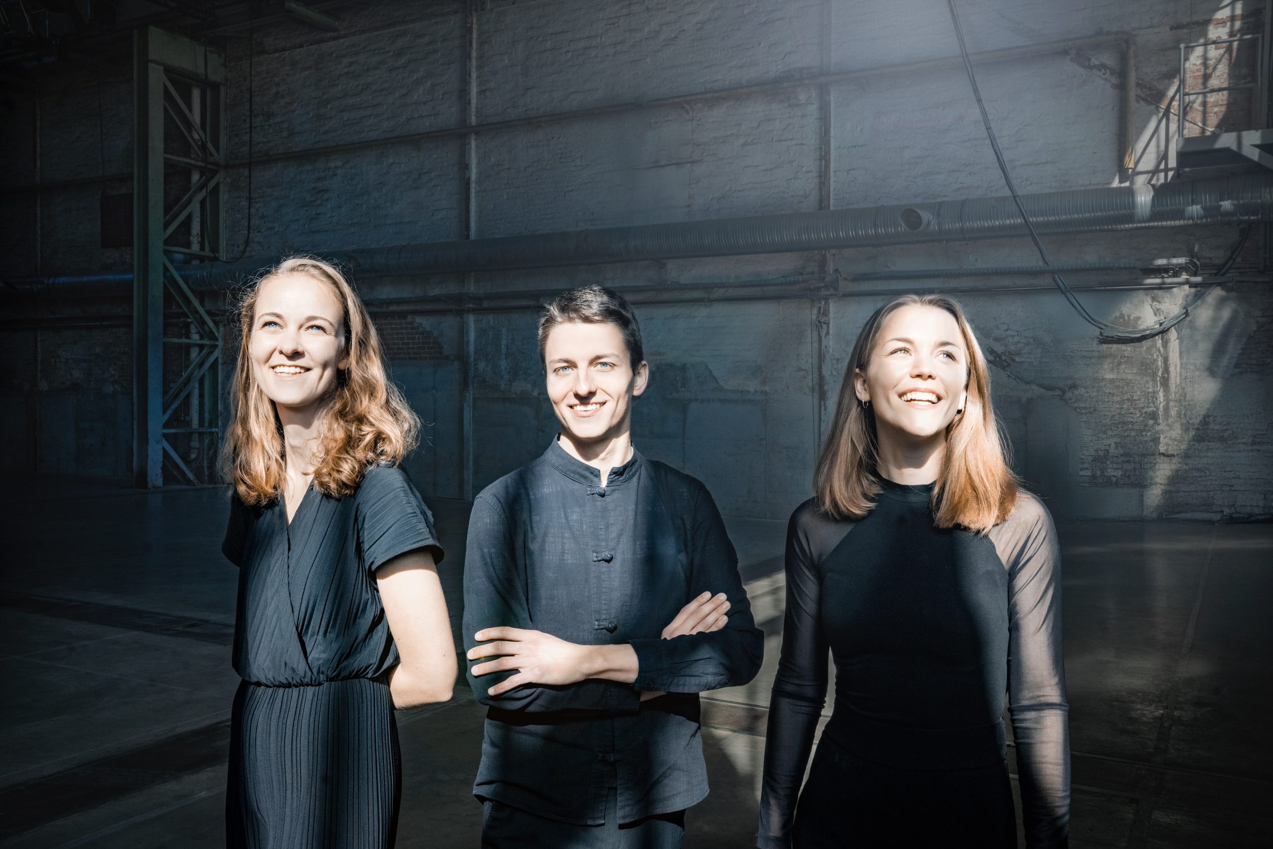 amelio trio - IN München - Das Stadtmagazin Three young adults in dark clothing smile in a dim, industrial warehouse with exposed pipes and brick walls behind them.