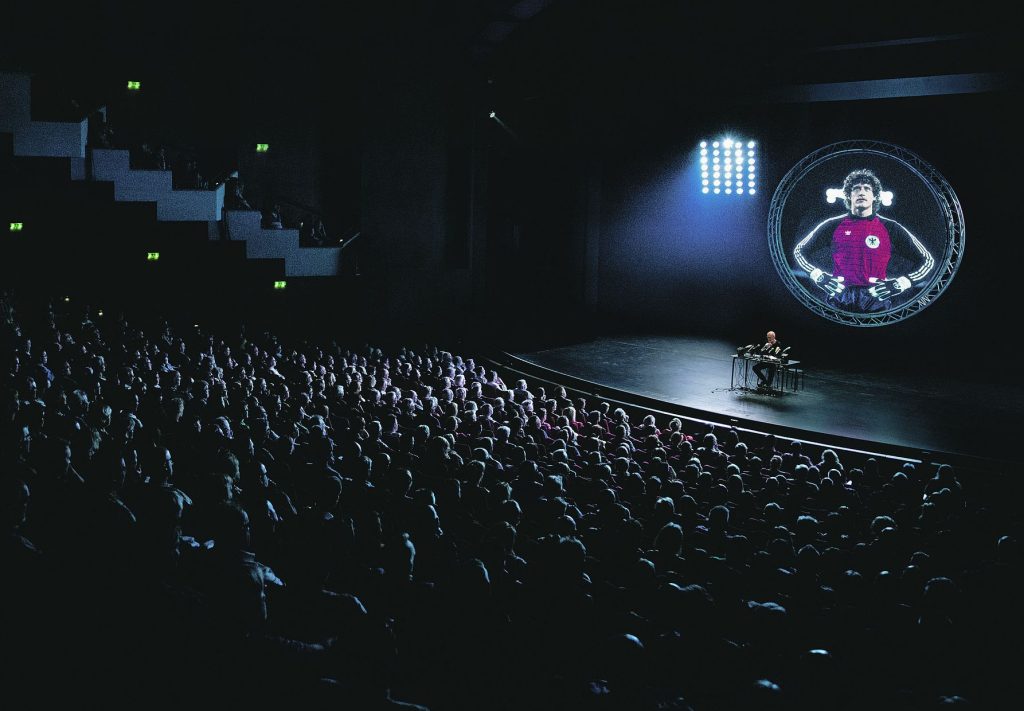 Presenter at a desk on a dark stage, facing a large audience; a circular screen behind them shows a person in a red jersey.