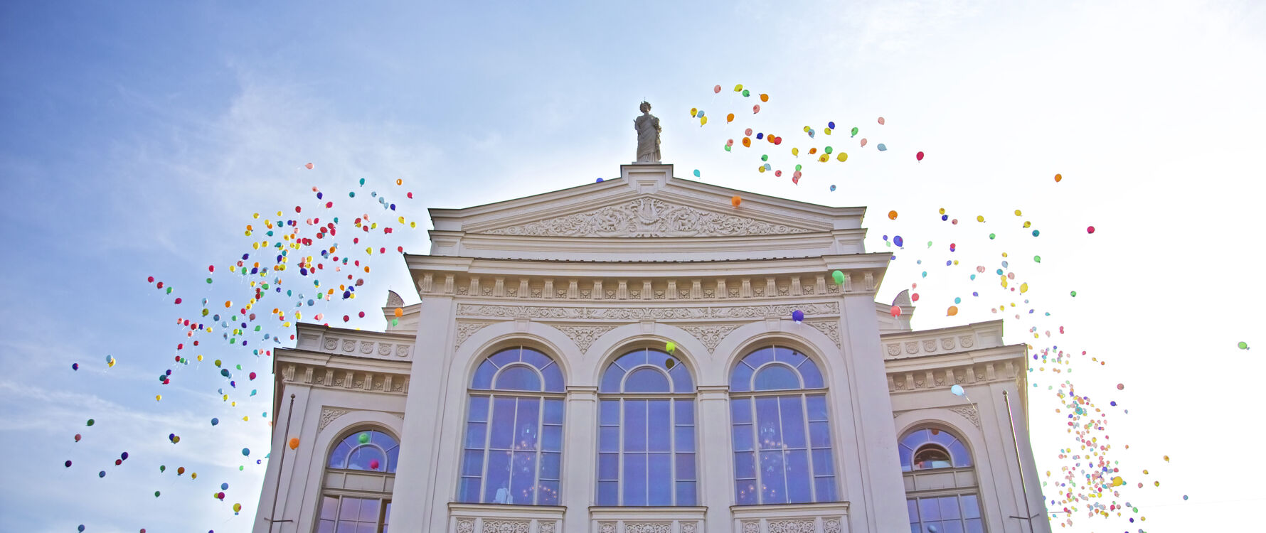 gpt-opening1819-christianpogozach-1 - IN München - Das Stadtmagazin Elegant white building with a statue on top and colorful balloons rising into a blue sky, suggesting celebration.
