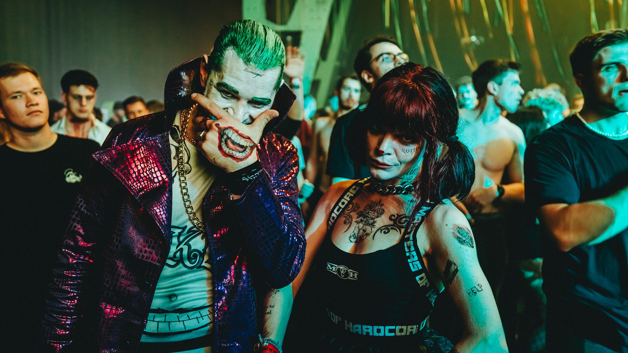 harderhorrornight_Kesselhaus_Halloween_2026 - IN München - Das Stadtmagazin Man in a shiny purple jacket with Joker-style grin makeup and a gold chain covers part of his face with his hand, standing beside a tattooed woman at a crowded club.