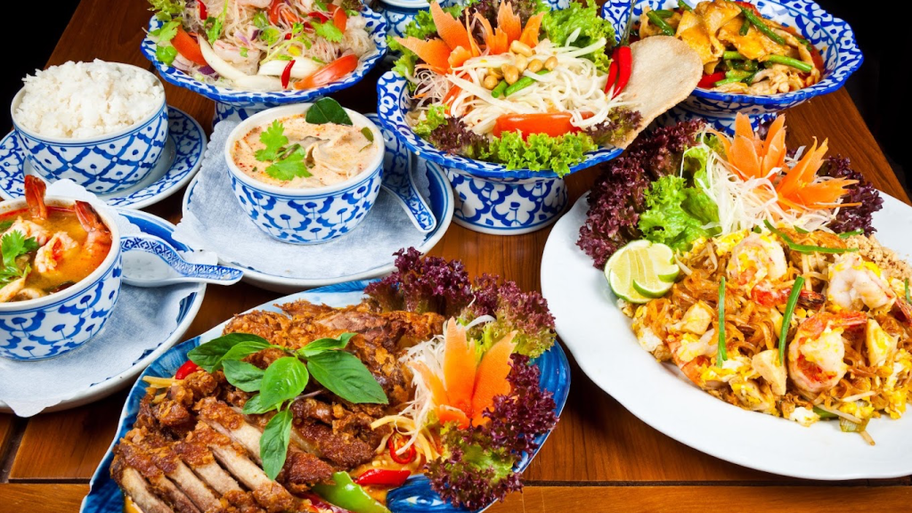 image - IN München - Das Stadtmagazin Assorted Thai dishes on a wooden table: fried meat, noodles, curry, rice, and fresh salad in blue-and-white bowls.