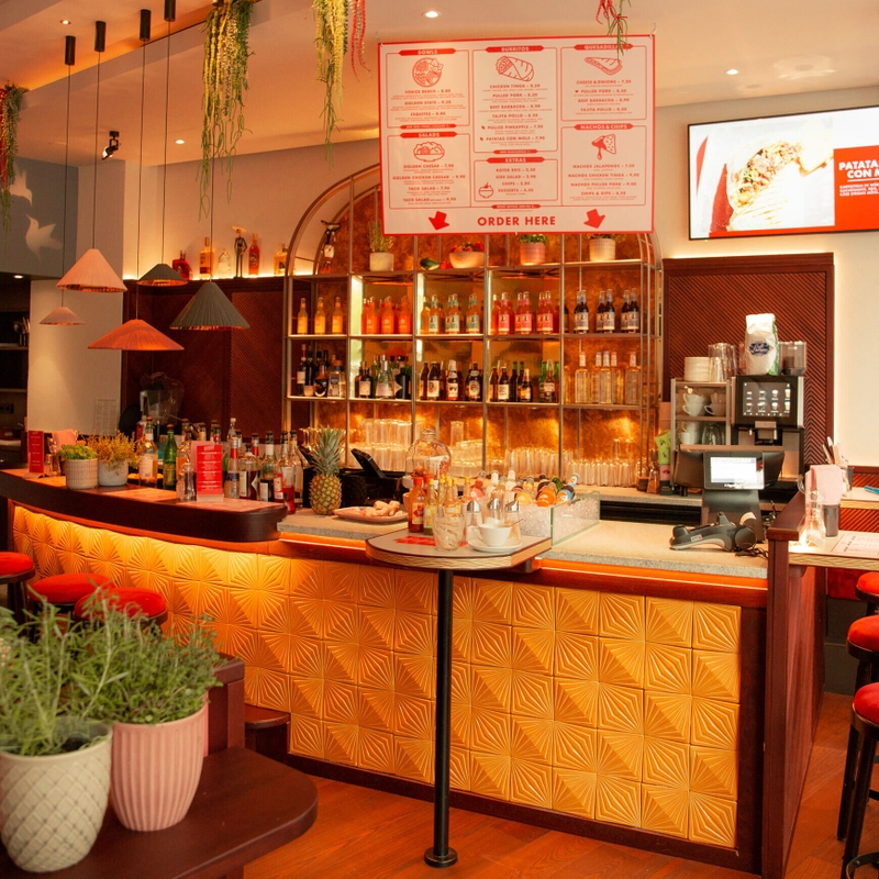 image - IN München - Das Stadtmagazin Bar area with backlit shelves of bottles, a red-framed menu board overhead, and a patterned gold front panel on the counter.