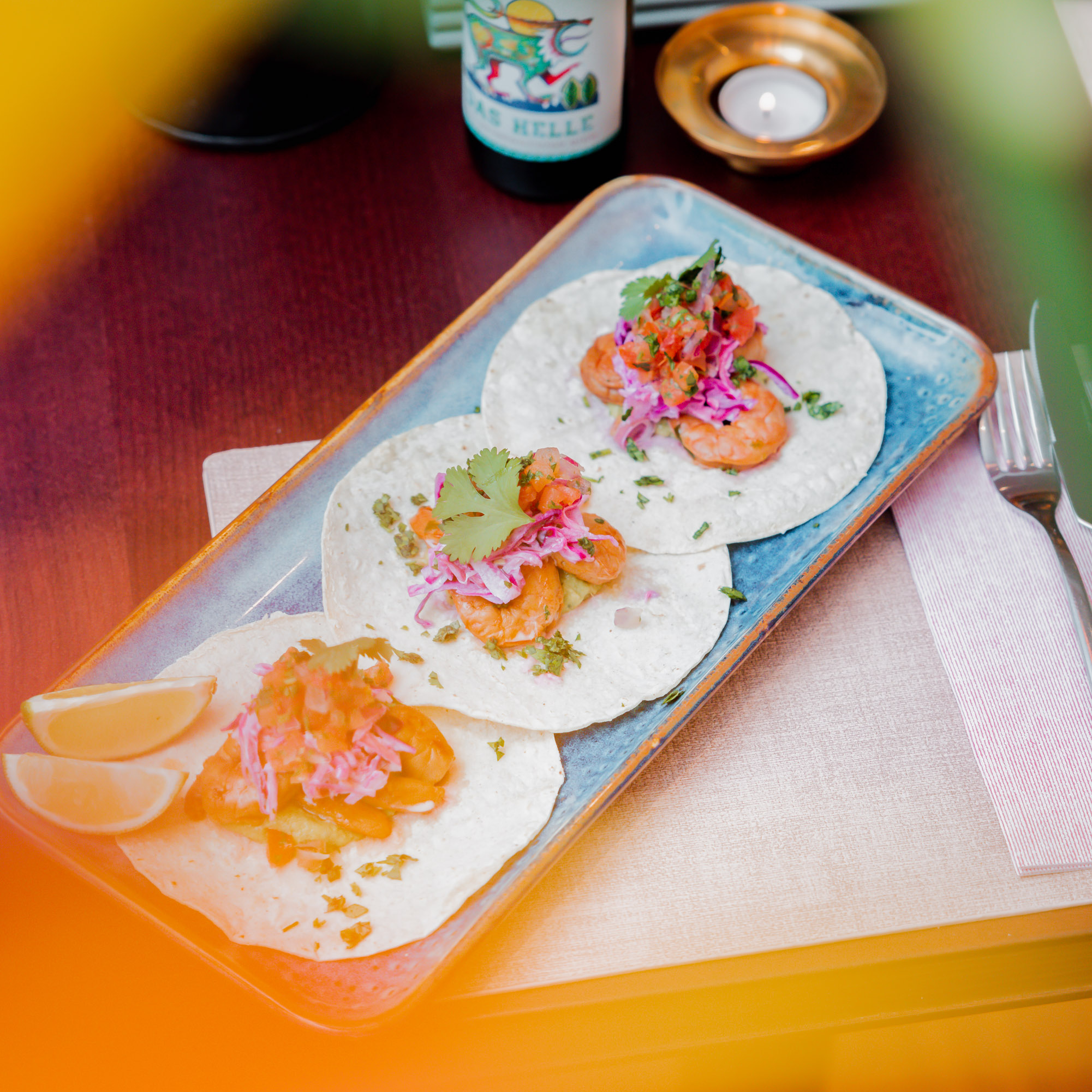 image - IN München - Das Stadtmagazin Shrimp tacos on a blue plate topped with pico de gallo, cilantro, and lime wedges.