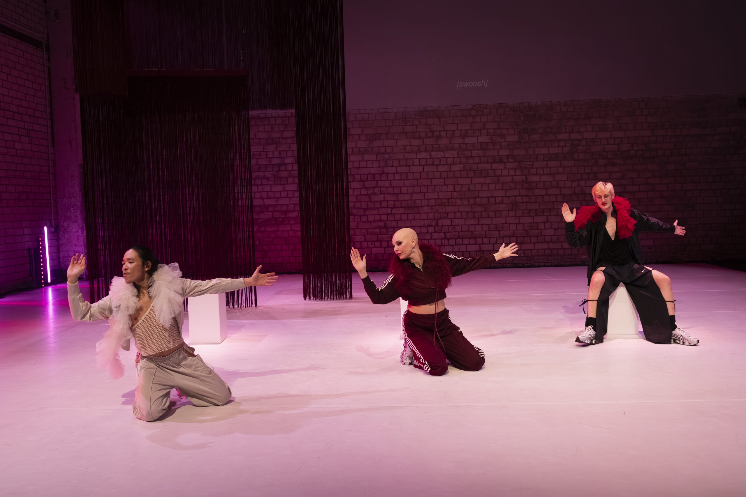 Contemporary dancers mid-performance on a pink-lit stage, with outstretched arms and bold costumes.