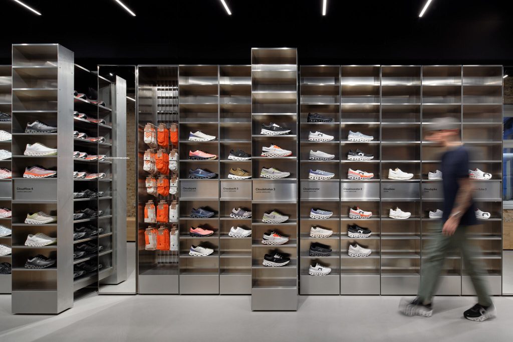 on_store-dtc-de-berlin-mar-2024-02 - IN München - Das Stadtmagazin Interior of a modern sneaker store with tall metal shelves holding rows of athletic shoes; a blurred shopper passes by on the right.