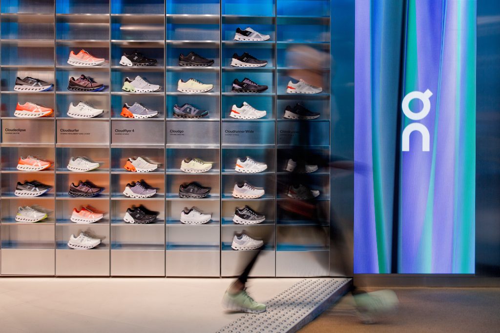 on_store-dtc-fr-paris-st_germain-nov-2024-03 - IN München - Das Stadtmagazin Shelf of running shoes on metal display panels in a store, with a blurred passerby moving past.
