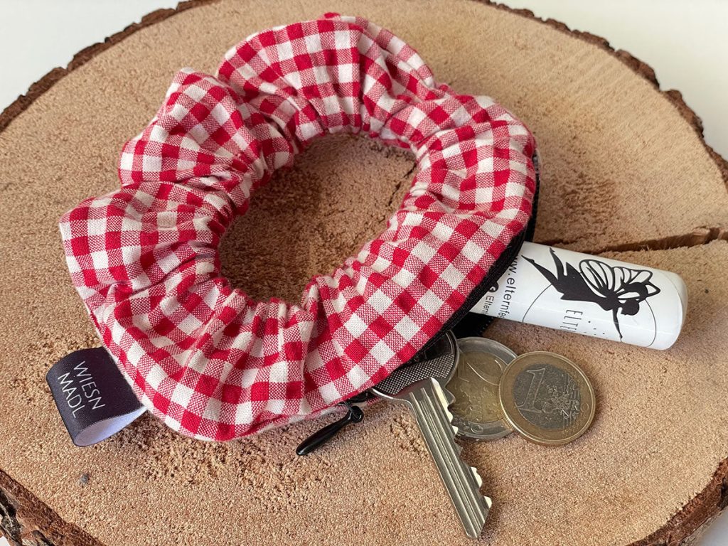 Red gingham scrunchie wrapped around a keyring with keys, a rolled receipt, and coins on a wooden tree-slice surface.