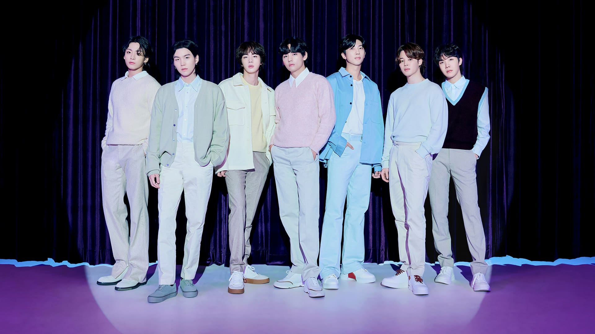 Eight young men stand side by side on a stage with a purple curtain backdrop, wearing pastel-colored outfits.