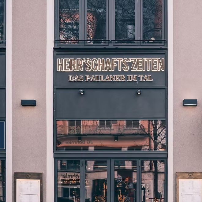 Building entrance with a large sign reading 'HERRSCHAFTSZEITEN' and 'DAS PAULANER IM TAL' above glass doors, reflections of trees visible in the windows.