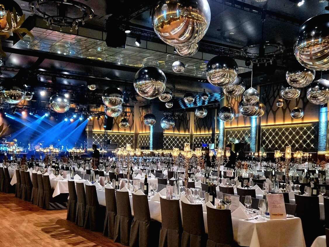 608665067_18393247084180572_3386568805656336637_n - IN München - Das Stadtmagazin Elegant banquet hall prepared for a formal event with long rows of tables, white linens, and black chair covers beneath hanging disco balls.